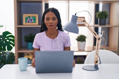 Photo for African american woman with braids using laptop at home scared and amazed with open mouth for surprise, disbelief face - Royalty Free Image