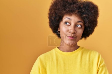 Foto de Joven mujer afroamericana de pie sobre fondo amarillo sonriendo mirando hacia un lado y mirando hacia otro pensando. - Imagen libre de derechos