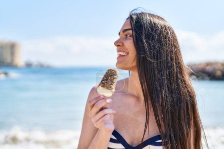Foto de Young hispanic woman wearing bikini eating ice cream at seaside - Imagen libre de derechos
