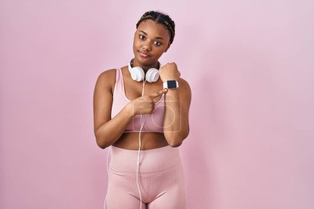 Téléchargez les photos : Femme afro-américaine avec des tresses portant des vêtements de sport et des écouteurs pressés pointant vers regarder le temps, l'impatience, regardant la caméra avec une expression détendue - en image libre de droit