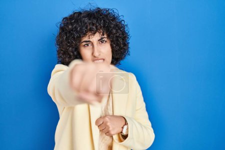 Photo for Young brunette woman with curly hair standing over blue background punching fist to fight, aggressive and angry attack, threat and violence - Royalty Free Image