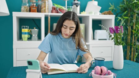 Photo for Young beautiful hispanic woman reading book sitting on table at dinning room - Royalty Free Image