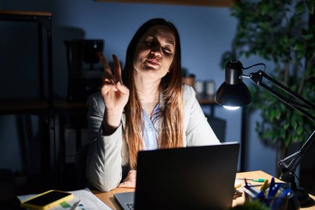 Photo for Young brunette woman working at the office at night showing and pointing up with fingers number two while smiling confident and happy. - Royalty Free Image