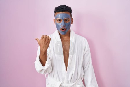 Photo for Young hispanic man wearing beauty face mask and bath robe surprised pointing with hand finger to the side, open mouth amazed expression. - Royalty Free Image