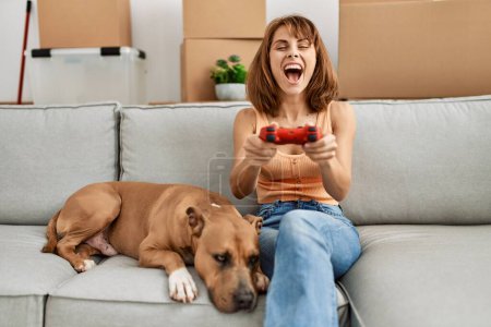 Téléchargez les photos : Jeune femme caucasienne jouant jeu vidéo assis sur le canapé avec chien à la maison - en image libre de droit