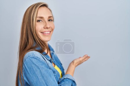 Photo for Young blonde woman standing over blue background inviting to enter smiling natural with open hand - Royalty Free Image