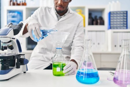 Téléchargez les photos : Jeune homme afro-américain portant un uniforme scientifique mesurant le liquide au laboratoire - en image libre de droit