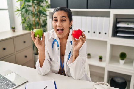 Photo for Young hispanic woman working at dietitian clinic holding green apple celebrating crazy and amazed for success with open eyes screaming excited. - Royalty Free Image