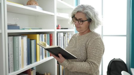 Foto de Middle age woman with grey hair teacher reading book at university classroom - Imagen libre de derechos