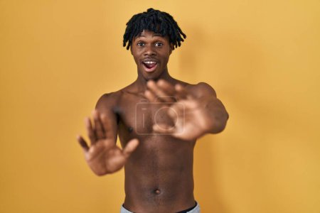 Téléchargez les photos : Jeune homme africain avec dreadlocks debout torse nu effrayé et terrifié par l'expression de la peur arrêter geste avec les mains, criant sous le choc. concept de panique. - en image libre de droit