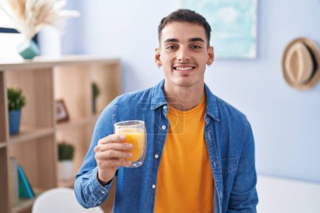 Foto de Hombre hispano guapo bebiendo un vaso de jugo de naranja luciendo positivo y feliz de pie y sonriendo con una sonrisa confiada mostrando los dientes - Imagen libre de derechos