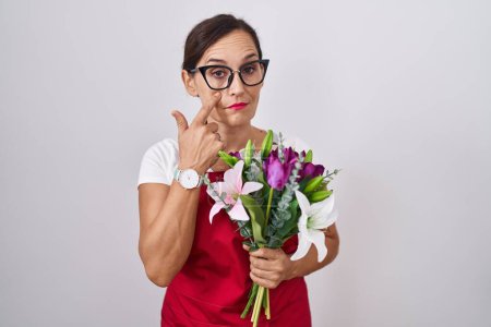 Photo for Middle age brunette woman wearing apron working at florist shop holding bouquet pointing to the eye watching you gesture, suspicious expression - Royalty Free Image