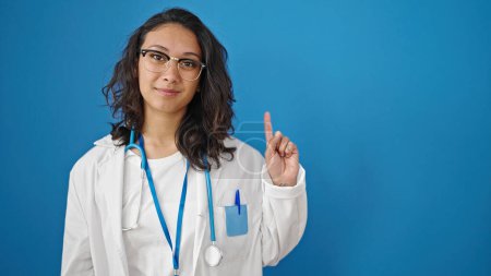 Téléchargez les photos : Jeune belle femme hispanique médecin souriant confiant debout avec le doigt levé sur fond de mur bleu isolé - en image libre de droit