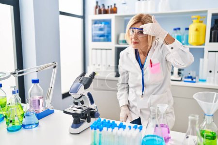 Photo for Middle age blonde woman working at scientist laboratory pointing unhappy to pimple on forehead, ugly infection of blackhead. acne and skin problem - Royalty Free Image