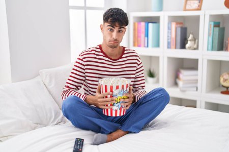 Photo for Young hispanic man eating popcorn sitting on the bed watching a movie skeptic and nervous, frowning upset because of problem. negative person. - Royalty Free Image