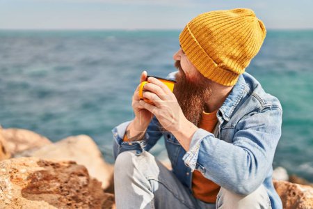 Foto de Young redhead man smiling confident drinking cup of coffee at seaside - Imagen libre de derechos