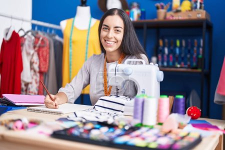 Photo for Young beautiful hispanic woman tailor smiling confident drawing on notebook at clothing factory - Royalty Free Image