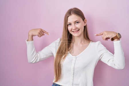 Téléchargez les photos : Jeune femme caucasienne debout sur fond rose regardant confiant avec le sourire sur le visage, se montrant du doigt fier et heureux. - en image libre de droit