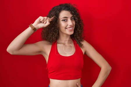 Photo for Hispanic woman with curly hair standing over red background smiling pointing to head with one finger, great idea or thought, good memory - Royalty Free Image