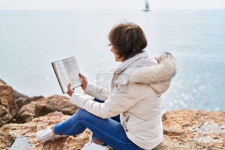 Photo for Middle age woman reading book sitting on the rock at seaside - Royalty Free Image