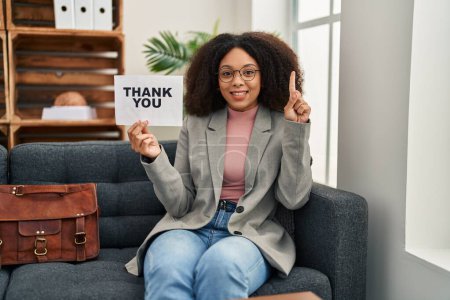 Foto de Mujer afroamericana joven que trabaja en la oficina de consulta sosteniendo banner de agradecimiento sonriendo con una idea o pregunta señalando el dedo con la cara feliz, número uno - Imagen libre de derechos