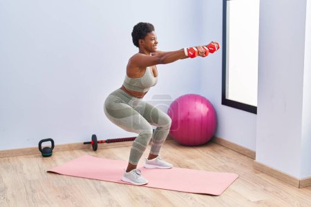 Téléchargez les photos : Femme afro-américaine souriant jambes d'entraînement confiantes exerce à l'aide d'haltères au centre sportif - en image libre de droit