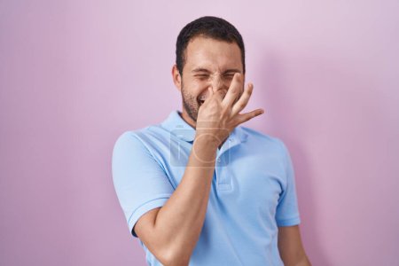 Foto de Hombre hispano de pie sobre fondo rosa oliendo algo apestoso y asqueroso, olor intolerable, conteniendo la respiración con los dedos en la nariz. mal olor - Imagen libre de derechos