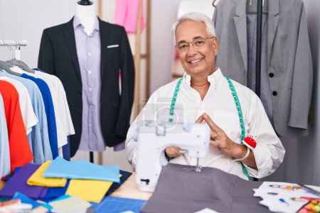 Photo for Middle age man with grey hair dressmaker using sewing machine hands together and fingers crossed smiling relaxed and cheerful. success and optimistic - Royalty Free Image