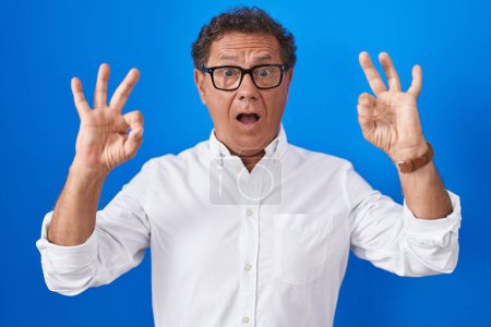Photo for Middle age hispanic man standing over blue background looking surprised and shocked doing ok approval symbol with fingers. crazy expression - Royalty Free Image