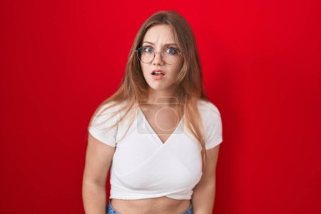 Photo for Young caucasian woman standing over red background in shock face, looking skeptical and sarcastic, surprised with open mouth - Royalty Free Image