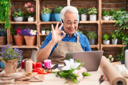 Foto de Hombre de mediana edad con el pelo gris trabajando en floristería haciendo videollamada haciendo signo bien con los dedos, sonriendo gesto amistoso excelente símbolo - Imagen libre de derechos