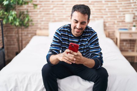 Photo for Young hispanic man using smartphone sitting on bed at bedroom - Royalty Free Image