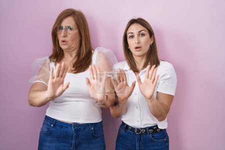 Photo for Hispanic mother and daughter wearing casual white t shirt over pink background moving away hands palms showing refusal and denial with afraid and disgusting expression. stop and forbidden. - Royalty Free Image