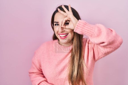 Téléchargez les photos : Jeune femme hispanique debout sur fond rose faisant un geste correct avec la main souriante, les yeux regardant à travers les doigts avec un visage heureux. - en image libre de droit