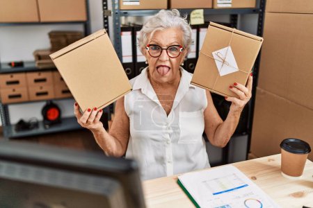Foto de Mujer mayor con cabello gris trabajando en el comercio electrónico de pequeñas empresas sacando la lengua feliz con expresión divertida. - Imagen libre de derechos