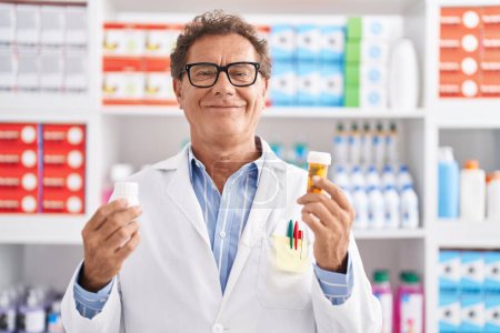 Téléchargez les photos : Homme moyen pharmacien souriant confiant tenant bouteilles pilules à la pharmacie - en image libre de droit