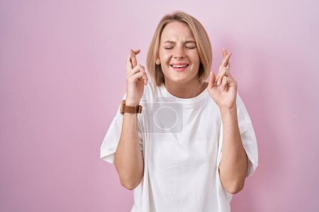 Téléchargez les photos : Jeune femme caucasienne debout sur fond rose geste doigt croisé souriant avec espoir et les yeux fermés. chance et concept superstitieux. - en image libre de droit
