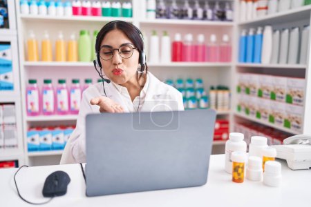 Téléchargez les photos : Jeune femme arabe travaillant à la pharmacie pharmacie en utilisant un ordinateur portable en regardant la caméra souffler un baiser avec la main sur l'air étant belle et sexy. expression d'amour. - en image libre de droit