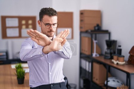Joven hombre hispano en la oficina expresión de rechazo cruzando brazos y palmas haciendo signo negativo, cara enojada 