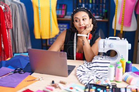 Photo for Young beautiful hispanic woman tailor using sewing machine and laptop at clothing factory - Royalty Free Image