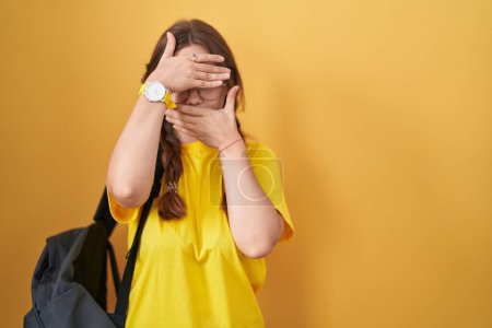 Foto de Mujer caucásica joven con mochila de estudiante sobre fondo amarillo que cubre los ojos y la boca con las manos, sorprendida y conmocionada. ocultar emociones - Imagen libre de derechos