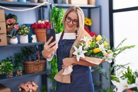Téléchargez les photos : Jeune femme blonde fleuriste faire selfie par smartphone tenant bouquet de fleurs à la boutique de fleuristes - en image libre de droit