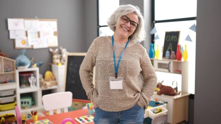 Téléchargez les photos : Middle age woman with grey hair preschool teacher smiling confident standing at kindergarten - en image libre de droit