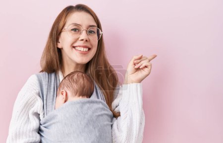 Foto de Joven mujer caucásica sosteniendo y llevando al bebé en un cabestrillo sonriendo feliz señalando con la mano y el dedo hacia un lado - Imagen libre de derechos