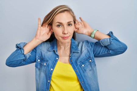 Téléchargez les photos : Jeune femme blonde debout sur fond bleu essayant d'entendre les deux mains sur le geste de l'oreille, curieuse de commérages. problème auditif, sourd - en image libre de droit