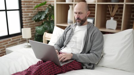 Téléchargez les photos : Jeune homme chauve utilisant un ordinateur portable assis sur le lit dans la chambre - en image libre de droit