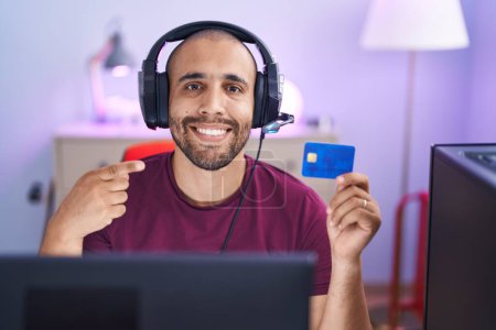Téléchargez les photos : Homme hispanique avec barbe faire des achats en ligne avec ordinateur et carte de crédit souriant heureux pointage avec la main et le doigt - en image libre de droit