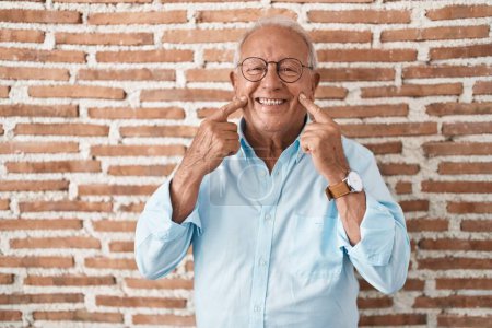 Téléchargez les photos : Homme âgé avec les cheveux gris debout sur le mur de briques souriant avec la bouche ouverte, les doigts pointant et forçant sourire joyeux - en image libre de droit