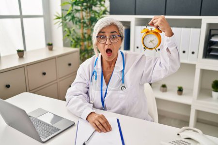 Foto de Mujer de mediana edad con pelo gris vistiendo uniforme médico sosteniendo reloj despertador asustado y sorprendido con la boca abierta para sorpresa, cara de incredulidad - Imagen libre de derechos