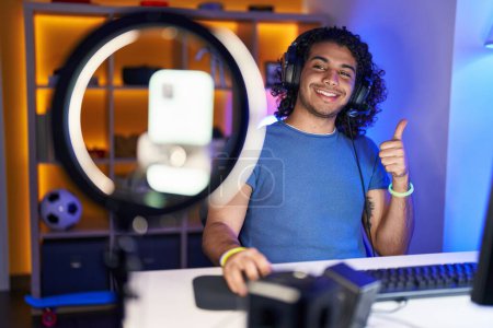 Foto de Hombre hispano con pelo rizado jugando videojuegos grabando con teléfono inteligente sonriendo feliz y positivo, pulgar hacia arriba haciendo excelente y signo de aprobación - Imagen libre de derechos